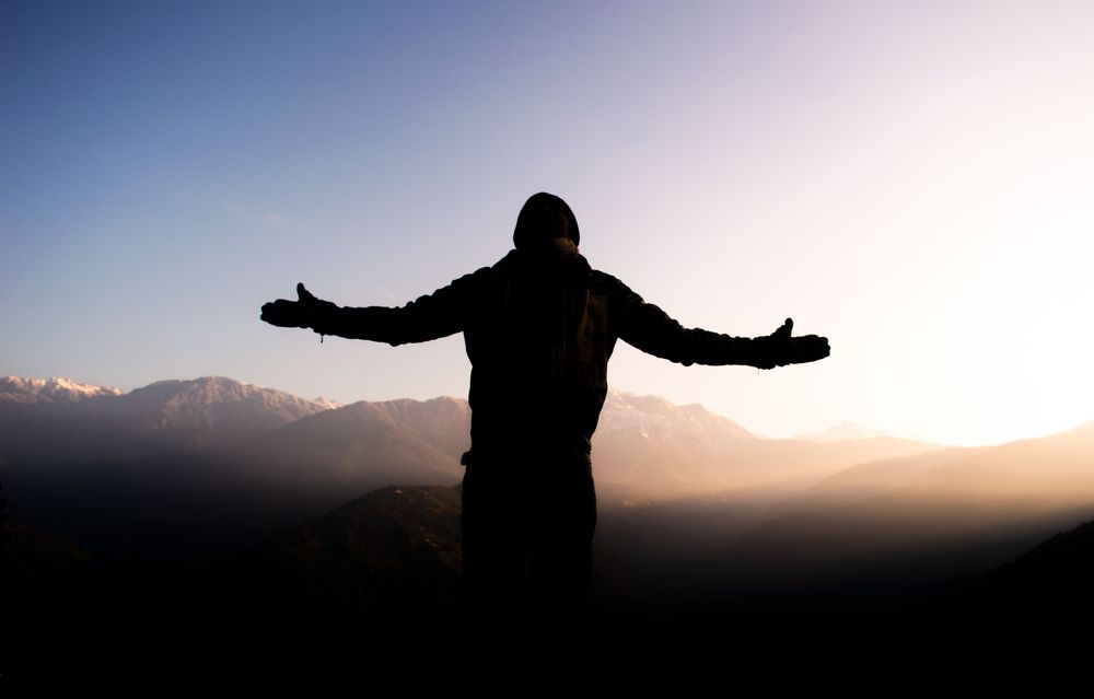 Une personne debout au sommet d'une montagne, bras ouverts face au lever du soleil
